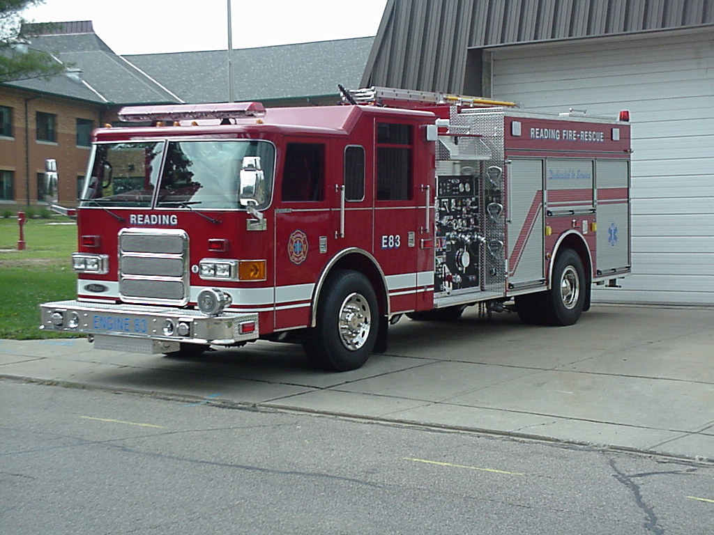 A red fire engine labeled "READING" is parked outside a building with a large garage door. It has various controls and equipment on its side, with "READING FIRE-RESCUE" written on the top. The vehicle is gleaming in the daylight.