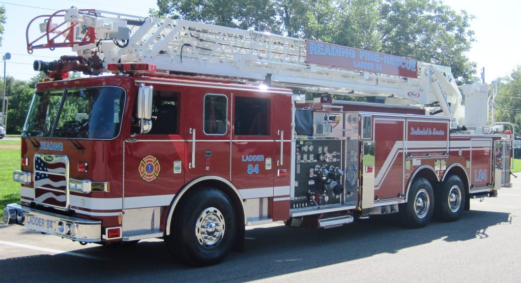 A red fire truck with "Reading Fire-Rescue" written on the side, featuring several compartments and an extended ladder on top. The truck is parked on a paved area, with trees and a sunny sky in the background.