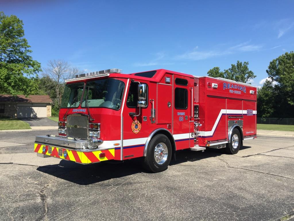 A bright red fire truck with the word "Reading" on the side is parked on a concrete surface. The truck features yellow and white striping. Trees and a building are visible in the background under a clear blue sky.