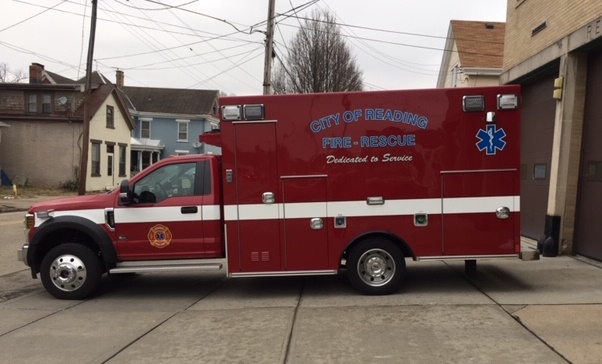A red ambulance labeled "City of Reading Fire-Rescue" is parked beside a building. The vehicle has white stripes and is marked with emergency symbols. Residential houses and power lines are visible in the background.