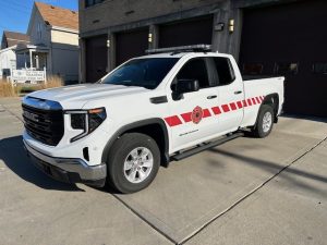 A white GMC pickup truck with red diagonal stripes and a fire department emblem on the door is parked outside a fire station on a sunny day.