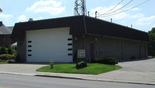 A one-story building with a large white garage door and brown roof. A sign and a fire hydrant are visible in front. Power lines and a communication tower are on top. Bushes and a road are in the foreground under a partly cloudy sky.
