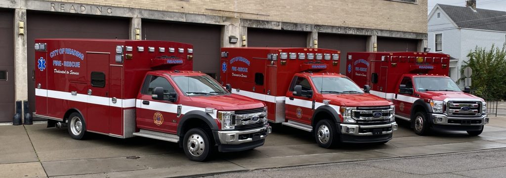 Three red and white emergency ambulances are parked in front of a beige brick fire station with large brown garage doors. The cloudy sky and wet pavement suggest recent rain. Sparse greenery and a neighboring house are visible.