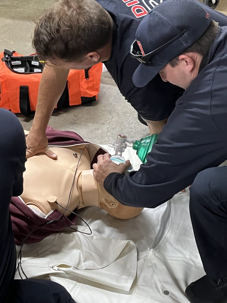 Two emergency responders practice CPR on a medical dummy. One performs chest compressions while the other uses a manual resuscitator. An orange equipment bag is visible in the background.