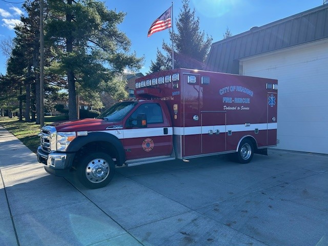 A red City of Hermitage fire rescue ambulance is parked outside a building with an American flag flying above. Trees and a blue sky are visible in the background.