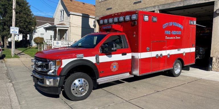 Red fire rescue ambulance parked outside a building in a residential area, with "City of Parma Fire - Rescue" written on the side. The vehicle has a white stripe and emergency lights on top. Nearby are houses and utility poles.