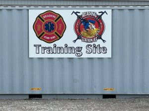 A sign on a metal wall reads "Reading Fire-EMS Training Site" with two emblems: one for Reading Fire-EMS, featuring a fire department logo, and another for Station 83, depicting a firefighter. Signs below say "Empty Only.