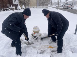 Two people wearing dark winter clothing shovel snow around a fire hydrant on a snowy street. Snow falls gently as they work, with houses and a car visible in the background.
