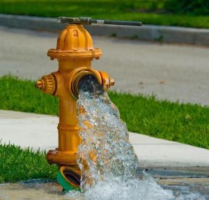 A yellow fire hydrant with its cap removed gushes water onto a sidewalk, with green grass and a paved street in the background.
