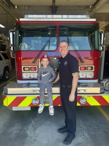 A smiling firefighter stands next to a young child who is sitting on the front bumper of a bright red fire truck inside a garage. The child is wearing a cap, glasses, a gray tracksuit, and sneakers.