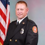 A firefighter in formal dress uniform poses for a portrait in front of an American flag. He is smiling and standing against a light-colored background.