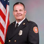 A male firefighter in formal dress uniform stands in front of an American flag, smiling, with a fire department badge and patch visible on his jacket.