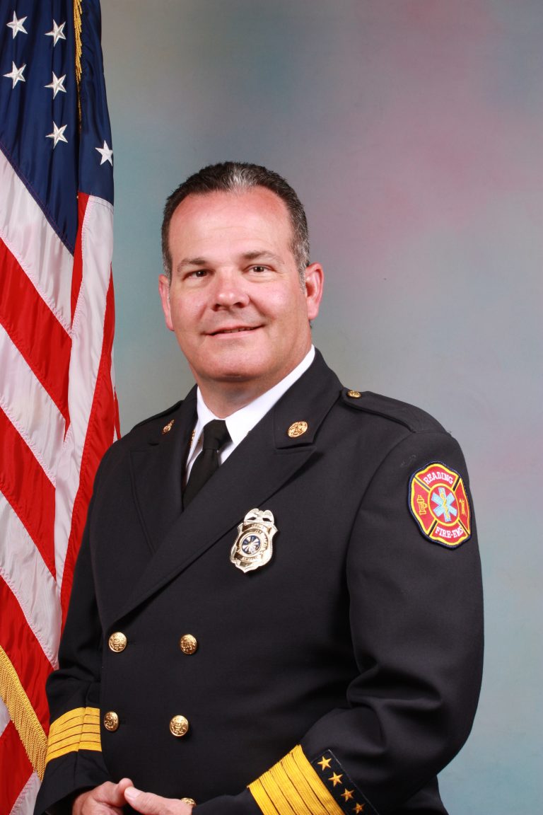 A male firefighter in formal dress uniform stands in front of an American flag, smiling, with a fire department badge and patch visible on his jacket.