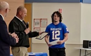 A man in khakis and a black jacket hands a certificate to a young person in a blue "Blue Devils 12" shirt during an indoor ceremony. Two other people stand nearby, and tables are set in the background.