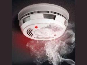 A smoke detector attached to a ceiling emits a red light as smoke wafts around it, indicating the presence of smoke or a potential fire. The device has ventilation slits and a central sensor area.
