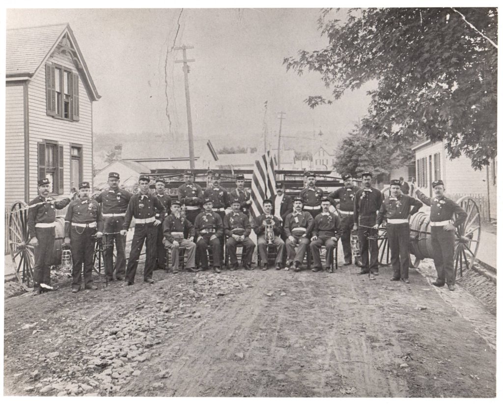 A vintage black-and-white photo of a group of uniformed men posing with musical instruments on a dirt road. They are flanked by wooden buildings and a horse-drawn carriage. An American flag is displayed in the center.