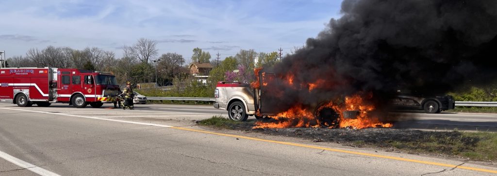 A white pickup truck is engulfed in flames and thick black smoke on the side of a highway. A fire truck and firefighters are nearby, responding to the emergency. The sky is partly cloudy.
