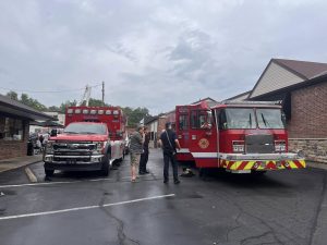 Two emergency vehicles—a fire truck and an ambulance—are parked in a lot outside brick buildings. Several people, including firefighters and civilians, are standing and talking near the vehicles.