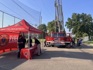 A Reading Fire Department event with firefighters at a booth under a red tent, a fire truck with an extended ladder nearby, and people gathered around in a sunny outdoor setting.