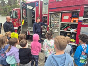 A firefighter explains equipment on a fire truck to a group of young children standing outside, listening attentively. Another firefighter stands nearby. The truck’s compartments with various tools are open and visible.