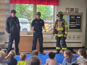 Three firefighters, one in full gear and two in uniforms, stand at the front of a classroom speaking to a group of young children seated on the floor. Alphabet posters and drawings decorate the classroom walls.