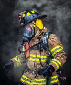 A firefighter wearing full protective gear, including a helmet, mask, and reflective suit, stands surrounded by smoke, ready for action in an emergency situation.