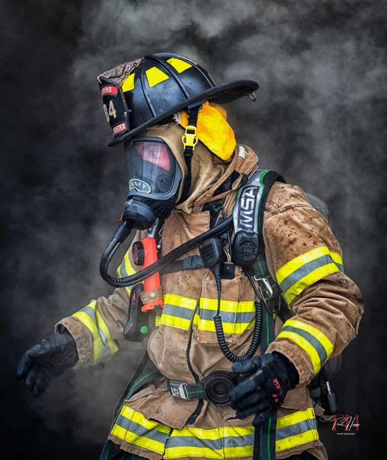 A firefighter wearing full protective gear, including a helmet, mask, and reflective suit, stands surrounded by smoke, ready for action in an emergency situation.