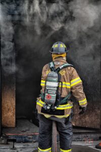 A firefighter wearing full gear and an oxygen tank stands in front of a smoke-filled doorway, preparing to enter a burning building.