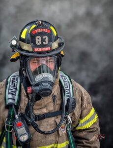 A firefighter in full gear, including a helmet labeled "Reading Firefighter 83" and a breathing mask, stands against a smoky background. The uniform is dirty, showing signs of recent use.