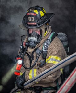A firefighter wearing full protective gear and an oxygen mask stands amid thick smoke, holding a hose and a flashlight. The helmet reads "83 Reading Firefighter." Reflective stripes are visible on the uniform.