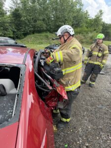A firefighter in protective gear uses hydraulic rescue tools to cut open the damaged door of a red car, while another firefighter stands nearby. The scene is outdoors, with trees and gravel in the background.