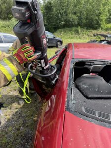 A firefighter uses hydraulic rescue tools to cut open the side of a red car with a shattered rear window during a vehicle extrication exercise.