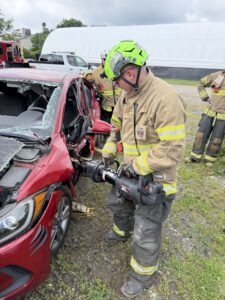 A firefighter in full gear uses a hydraulic rescue tool to cut open the door of a damaged red car during a training exercise. Other firefighters and emergency vehicles are visible in the background.