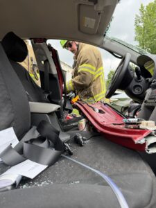 A firefighter in uniform stands outside a car with its door removed, likely during a rescue operation. The interior is messy, with shattered glass and seatbelts askew. The steering wheel and dashboard are visible.
