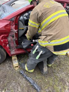 A firefighter uses rescue tools to pry open the door of a damaged red car during an emergency response drill. The car is stabilized with wooden blocks, and the firefighter is wearing safety gear and a helmet.