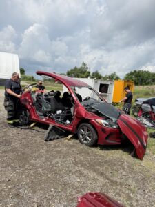 Three firefighters stand near a red car with heavy damage and its doors and roof removed, possibly during a rescue drill or accident simulation, with equipment scattered around and cloudy skies overhead.