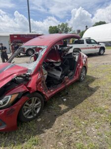 A heavily damaged red car with its roof partially torn off sits on grass. Emergency vehicles, including a fire truck and a white pickup, are parked nearby. The sky is partly cloudy.