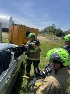Three firefighters in yellow gear and bright green helmets use hydraulic rescue tools to open the door of a silver car during a training exercise outdoors on a sunny day.