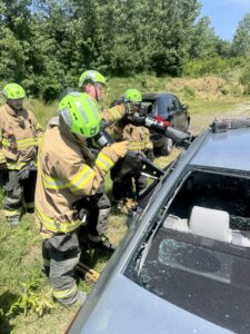 Four firefighters wearing yellow protective gear and green helmets use hydraulic rescue tools to cut through the roof of a silver car with shattered windows during a daytime training exercise outdoors.
