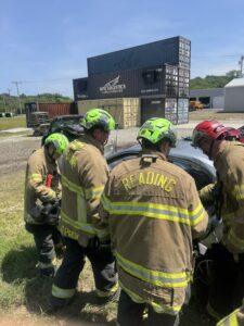 Four firefighters in yellow gear and green helmets, with "Reading" on their jackets, work together next to a car outdoors near stacked shipping containers and industrial buildings on a sunny day.