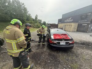 Three firefighters in gear stand next to a black sedan, using tools on the rear door. The car has a red tarp on the back window. Shipping containers and trees are visible in the background.