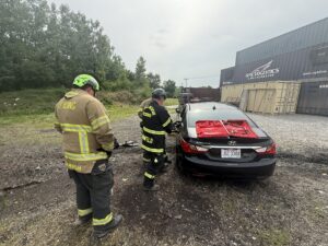 Two firefighters in gear stand near a black car with red equipment on its roof in a gravel lot, with shipping containers and trees in the background. One firefighter is writing on a clipboard.