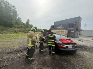 Three firefighters in protective gear stand beside a black car with red material on its back window, appearing to conduct a training exercise near shipping containers in an outdoor gravel area.
