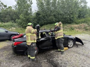 Two firefighters in protective gear use rescue tools to cut open the roof of a black car during an outdoor emergency response demonstration or training exercise. Trees and another vehicle are visible in the background.