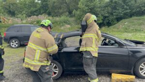 Two firefighters in gear and helmets use tools to open the door of a black car, likely during a rescue drill. Another parked vehicle and greenery are visible in the background. Yellow blocks are by the car’s front wheel.