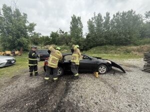 Three firefighters in full gear practice a vehicle rescue, working on a black car with its hood open in a gravel lot surrounded by trees and equipment.