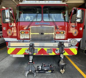 A red fire truck with "READING" on the front is parked in a fire station. Its cab doors are open, and hydraulic rescue tools (Jaws of Life) are positioned in front of the truck on the concrete floor.
