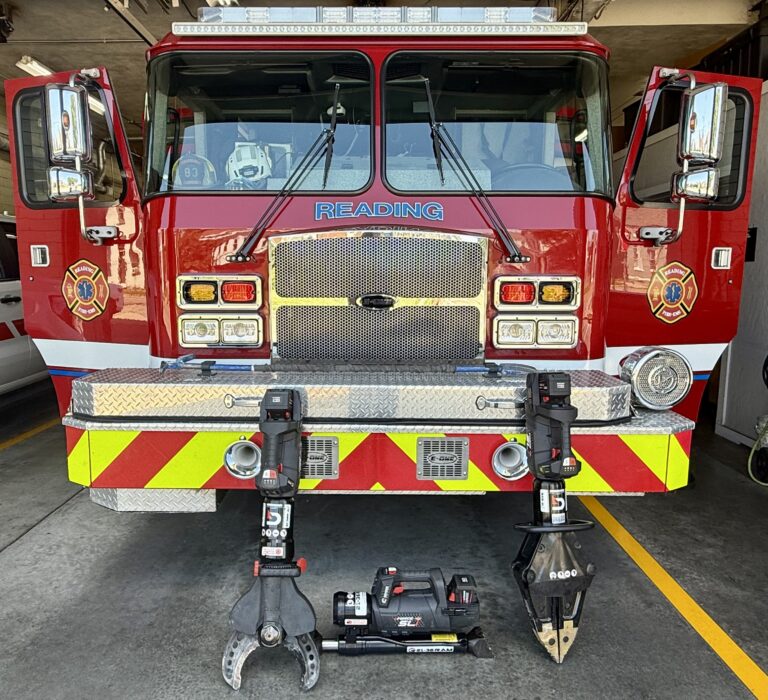 A red fire truck with "READING" on the front is parked in a fire station. Its cab doors are open, and hydraulic rescue tools (Jaws of Life) are positioned in front of the truck on the concrete floor.