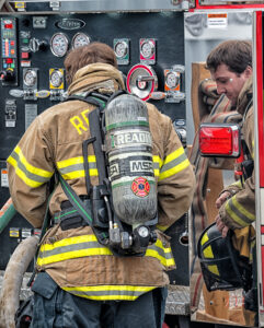 Two firefighters in full gear stand near a fire truck's control panel. One adjusts controls, while the other, holding a helmet, is reflected in a shiny surface. Safety equipment and gauges are visible.