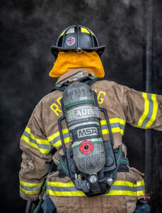 A firefighter in full gear, including a helmet and oxygen tank labeled "READING MSA," stands with their back to the camera, facing a smoky, dark environment.